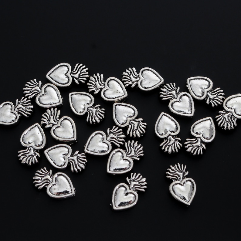 May include: A collection of silver-toned heart-shaped charms with a flame design. The charms are scattered on a black background.