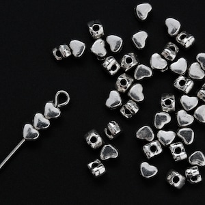 May include: A collection of silver heart-shaped beads, some strung on a wire, others loose. The beads are perfect for jewelry making or crafting.