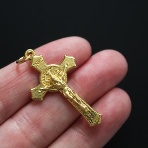 Op de afbeelding: Een goudkleurige kruisbeeldhanger. Het kruis heeft een gedetailleerde afbeelding van Jezus Christus en de letters "INRI" bovenaan. De hanger wordt tegen een donkere achtergrond gehouden.