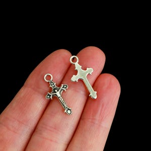 May include: Two silver-toned metal cross charms. One charm features a detailed embossed figure of a person on the cross, while the other charm is a plain, smooth cross shape. Both pieces include a small circular loop at the top for attachment.