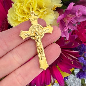 Op de afbeelding: Een goudkleurige kruisbeeldhanger met de figuur van Jezus Christus. Het kruis heeft de letters INRI bovenaan. De hanger wordt vastgehouden voor een kleurrijk bloemstuk met gele, roze en paarse bloemen.