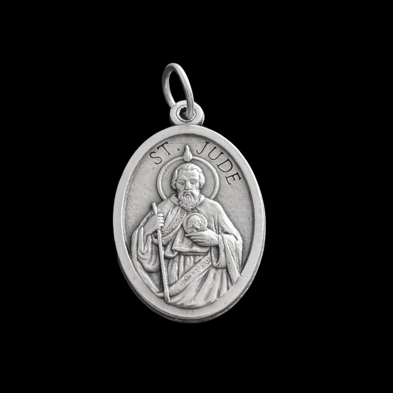 Op de afbeelding: Zilveren ovale hanger met een afbeelding van Sint Judas Thaddeus, beschermheilige van hopeloze gevallen. De hanger toont een gedetailleerde afbeelding van Sint Judas die een staf vasthoudt en een vlam boven zijn hoofd. De inscriptie "ST. JUDE" is gegraveerd rond de rand van de hanger.