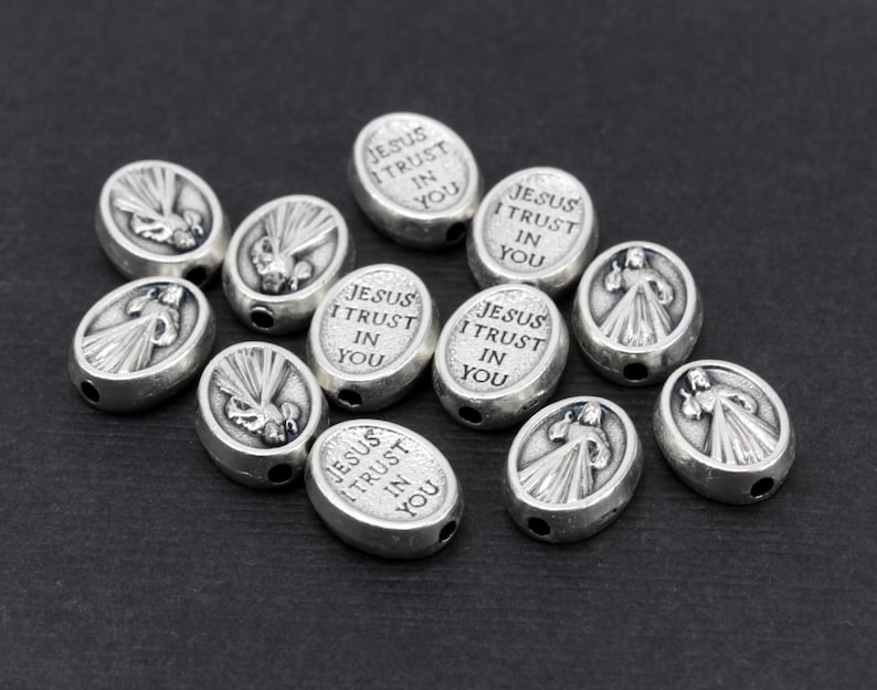 Puede incluir: Cuentas ovaladas de plata con las palabras "Jes&uacute;s, conf&iacute;o en ti" grabadas. Las cuentas presentan una imagen en relieve de una paloma con rayos de luz.