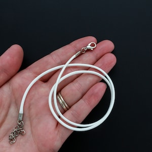 Puede incluir: Collar de cordón blanco con cierre plateado y cadena extensora. El collar se muestra en una mano, colocado plano.