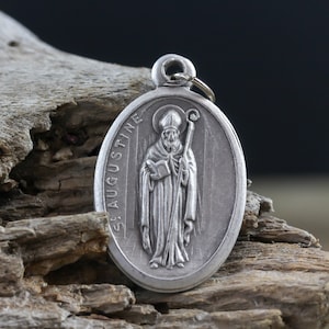 May include: Silver oval pendant with a depiction of Saint Augustine holding a book and a staff. The pendant has the words "St. Augustine" inscribed on the side.