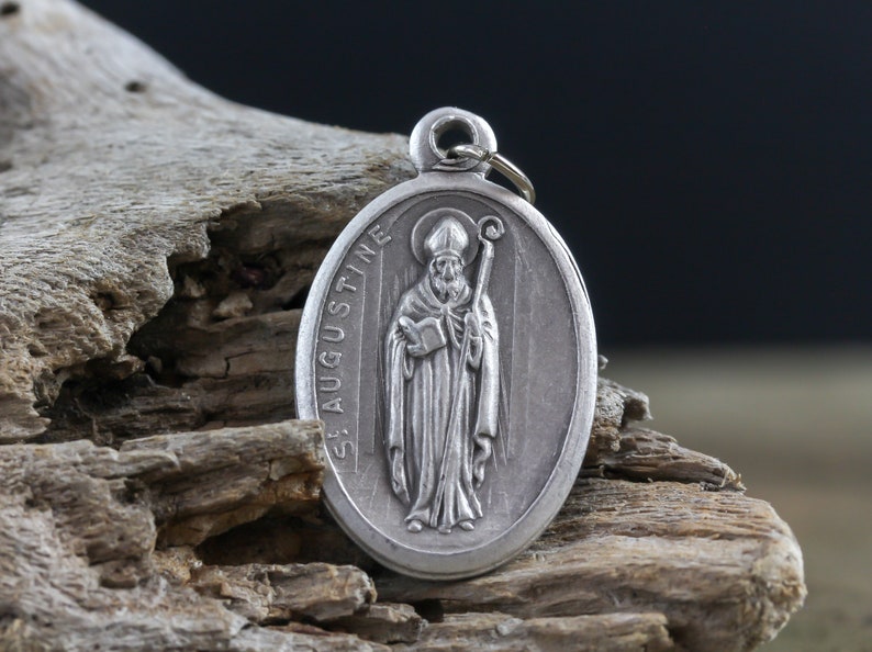 May include: Silver oval pendant with a depiction of Saint Augustine holding a book and a staff. The pendant has the words "St. Augustine" inscribed on the side.
