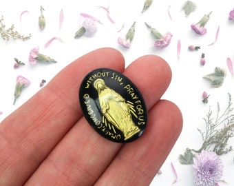 May include: A black oval charm with a gold image of the Virgin Mary. The charm has the text "Our Lady Conceived Without Sin, Pray For Us" around the edge.