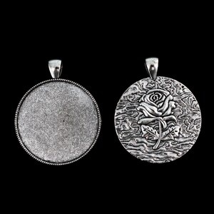 Pode incluir: Um pingente de prata com um design de rosa em um lado e uma superfície em branco no outro.