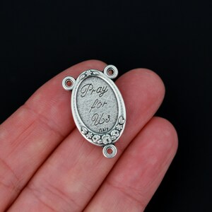 May include: Silver oval-shaped charm with the words "Pray for Us" and "Italy" engraved on it. The charm has a floral design around the edge and three jump rings for attaching to a necklace or bracelet.