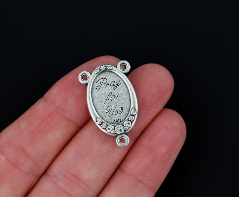 May include: Silver oval-shaped charm with the words "Pray for Us" and "Italy" engraved on it. The charm has a floral design around the edge and three jump rings for attaching to a necklace or bracelet.