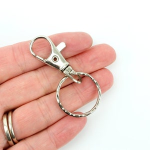 May include: A silver-coloured metal swivel clasp with a key ring. The clasp has a spring-loaded lever for easy attachment. The key ring is a textured, circular design. The clasp is shown against a white background.