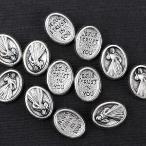 Puede incluir: Una colecci&oacute;n de dijes ovalados plateados con las palabras "Jes&uacute;s conf&iacute;o en ti" y una imagen de un coraz&oacute;n con rayos de luz.