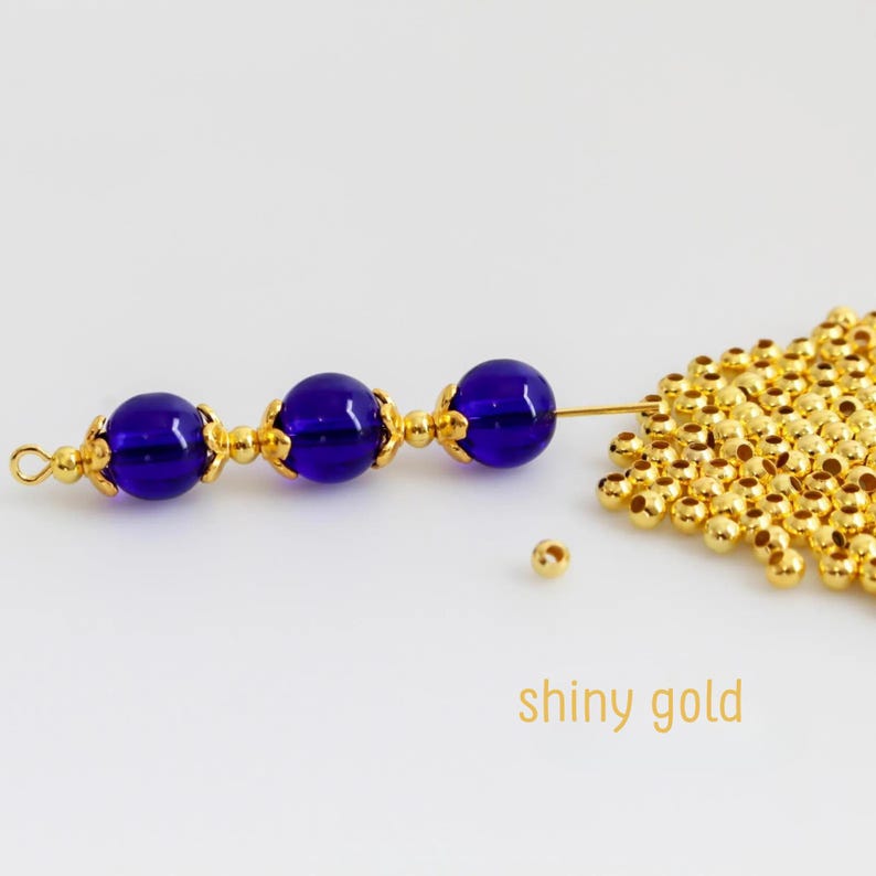May include: Three deep blue spherical beads separated by small gold-toned metal spacers on a thin wire, positioned next to a cluster of numerous tiny, shiny gold-toned spherical beads. The text "shiny gold" is printed in the lower right corner.