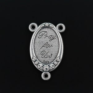May include: Silver oval charm with the words "Pray for Us" and "Italy" inscribed on it. The charm has a floral design around the edge and three loops for attaching to a chain or bracelet.