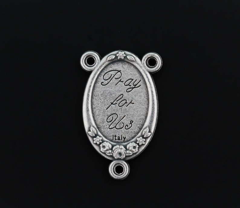 May include: Silver oval charm with the words "Pray for Us" and "Italy" inscribed on it. The charm has a floral design around the edge and three loops for attaching to a chain or bracelet.