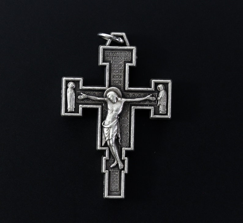 Op de afbeelding: Een zilverkleurige kruis hanger met een gedetailleerd ontwerp. Het kruis toont een figuur van Jezus Christus aan het kruis, met twee figuren aan weerszijden. De hanger heeft een lus aan de bovenkant om aan een ketting te bevestigen.