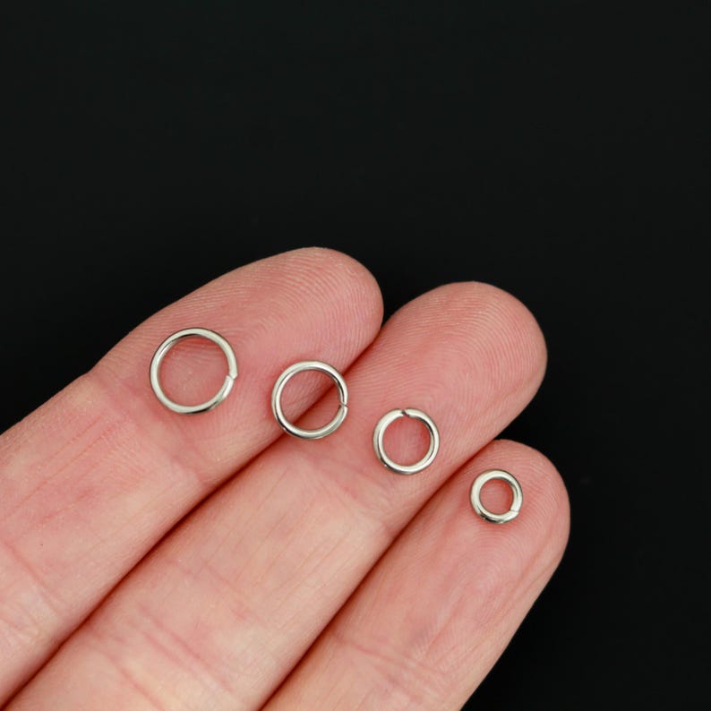 K&ouml;nnte beinhalten: Vier silberfarbene Metall-Biegeringe in verschiedenen Gr&ouml;&szlig;en, die in einer Reihe angeordnet sind. Jeder kreisf&ouml;rmige Ring weist eine kleine, deutliche &Ouml;ffnung entlang seines Umfangs auf, um Schmuckkomponenten oder andere kleine Bastelartikel sicher zu verbinden.