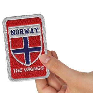 May include: A rectangular embroidered patch with a red background and a white border. It features a shield design with a blue cross on a white field, the text NORWAY in blue, and the text THE VIKINGS in white, all stitched with thread.
