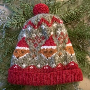 May include: A knit beanie featuring a grey base with a repeating pattern of orange fox faces wearing red hats. The hat has a solid red ribbed cuff and a matching red pom-pom on top. The design includes white accents and a zigzag border pattern.