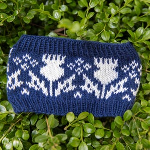 Op de afbeelding: Een donkerblauwe gebreide hoofdband met een wit fair isle-patroon van gestileerde bloemvormen en kleine geometrische accenten. Het accessoire heeft een geribbelde textuur langs de boven- en onderrand voor een consistent ontwerp.