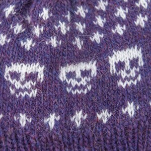 Close-up detail of a purple stranded colourwork knitted hat, showing the skull motif and small white cross stitches, highlighting the yarn texture and Fair Isle patterning.