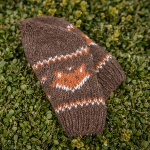 Side view of brown Fair Isle fox hat knitting pattern, showing fox motif band and ribbed brim, photographed on green foliage.
