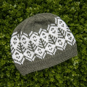 May include: A knit beanie in olive green featuring a wide band of white geometric patterns around the center. The hat is constructed with a ribbed brim at the base and a smooth stockinette stitch crown, showcasing a repeating diamond and tree motif.