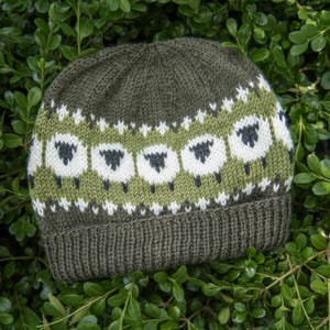May include: A dark olive green knit beanie featuring a horizontal band of white and dark grey sheep motifs. The hat has a ribbed cuff at the base and a solid dark olive green crown, showcasing a detailed fair isle pattern with small white accents.
