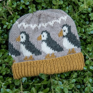 May include: A knit beanie featuring a repeating pattern of three puffin birds in black, white, and mustard yellow against a light brown background. The hat has a wide, ribbed mustard yellow cuff at the bottom and a white zigzag line above the birds.