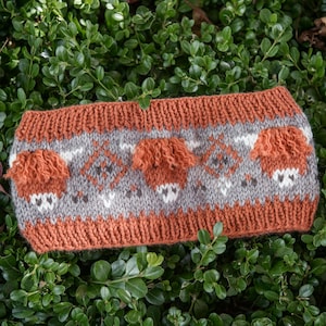 May include: A knitted headband featuring a repeating pattern of rust-colored Highland cattle with textured, fuzzy yarn details. The design includes gray and white geometric diamond motifs on a gray background, bordered by wide, solid rust-colored ribbed edges.