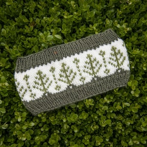 May include: A knitted headband featuring a wide band of white yarn decorated with a repeating pattern of olive green pine trees. The top and bottom edges of the headband are finished with a ribbed olive green knit border for a secure fit.