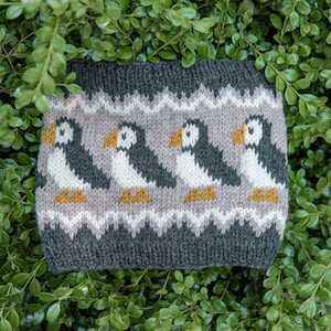 May include: A knitted headband featuring a repeating pattern of puffin birds. The design includes dark charcoal gray bands at the top and bottom, with a light gray center section displaying four puffins with white chests, black wings, and orange beaks.