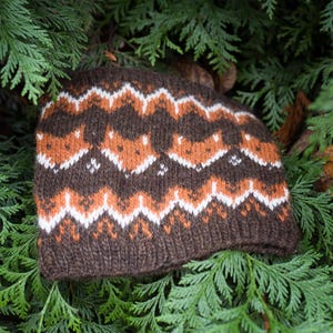 May include: A knitted beanie featuring a repeating pattern of stylized fox faces in orange, white, and dark brown. The hat has a ribbed hem at the base and a dense, textured knit construction with a consistent color palette throughout the design.
