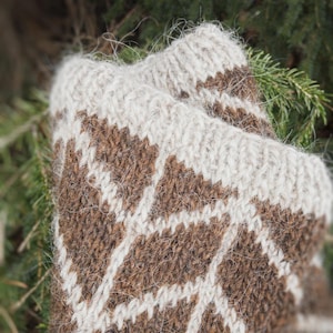May include: Close-up of a pair of knitted mittens. The mittens feature a brown and white geometric pattern. The cuffs are a solid off-white color. The mittens are made of a thick, textured yarn, suggesting warmth and comfort. The background is blurred.