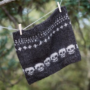Può includere: Scaldacollo lavorato a maglia grigio scuro con un motivo a teschio bianco e disegni geometrici. Lo scaldacollo è appeso a un filo con mollette di legno su uno sfondo verde sfocato. Un accessorio fatto a mano e accogliente.