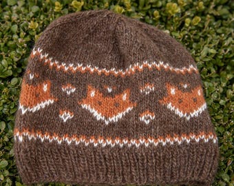 May include: A brown knit beanie featuring a repeating fair isle pattern of orange and white fox faces. The hat has a ribbed hem at the base and a solid brown crown, constructed with a textured yarn that displays visible knit stitches throughout.