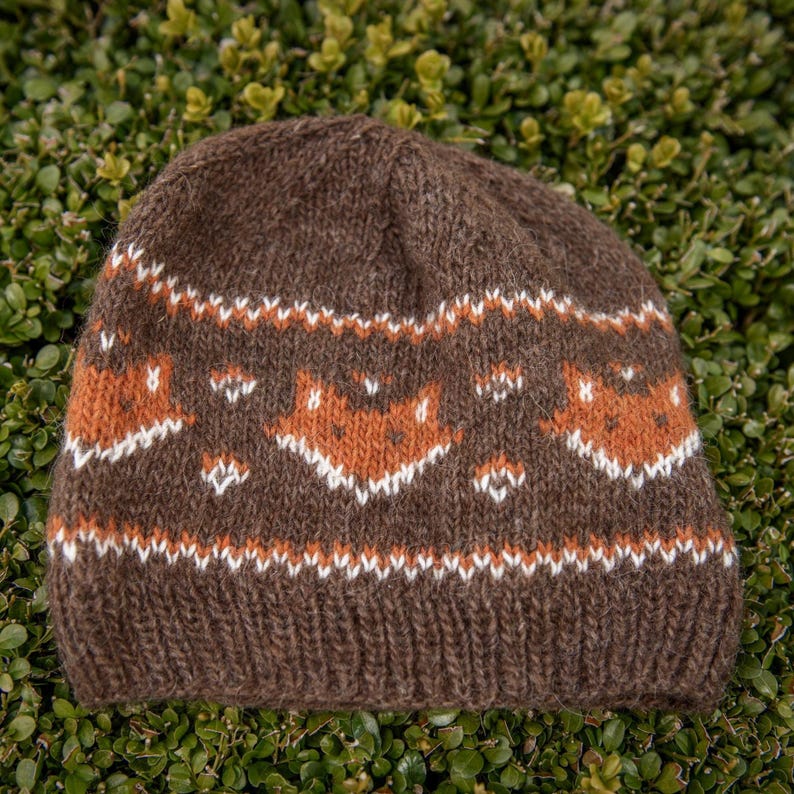 Brown Fair Isle fox hat knitting pattern with repeating fox motifs in rust and white, shown flat on greenery with ribbed brim.