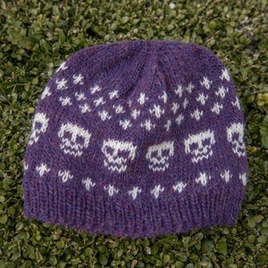 Purple stranded colourwork knitted hat laid on green foliage, featuring a repeating skull motif and small white cross details, shown flat to highlight the pattern and texture.