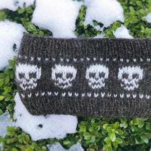 Op de afbeelding: Donkergrijze gebreide hoofdband met een herhalend patroon van witte schedels. Het schedelontwerp is gecentreerd op de hoofdband, met kleine witte stippen en chevrons boven en onder de schedels. De hoofdband ligt op groene bladeren met sneeuwvlekken.