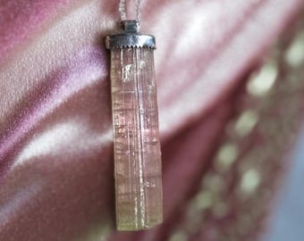 May include: A silver necklace featuring a rectangular, pink and clear tourmaline crystal pendant. The crystal has a natural, textured appearance and is suspended from a delicate silver chain. The pendant is approximately 1.5 inches long.