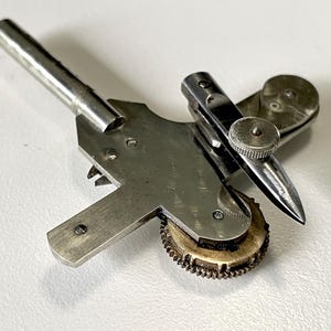May include: A silver-toned metal watchmaker's tool featuring a long cylindrical handle, a flat cross-shaped plate, a small circular gear with fine teeth, and an adjustable pointed arm secured by a knurled screw for precision mechanical work.