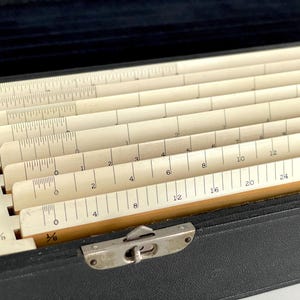 May include: An antique wooden slide rule set in a black case. The slide rules are cream-colored with black markings and measurements in inches. The case has a metal clasp and is open, revealing the stacked rulers.
