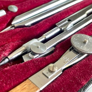 May include: Close-up of a vintage drafting set with silver-colored compasses and dividers. The tools are nestled in a red velvet-lined case. The compasses have a pencil and needle point. The set includes a small, silver-colored screw.