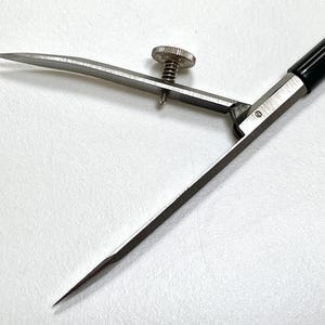 May include: A drafting compass featuring two sharp, pointed metal legs connected by a central hinge. One leg includes an adjustable silver-toned metal screw mechanism for precise positioning. The handle is a smooth, black cylindrical rod.