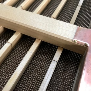 May include: Close-up of a wooden frame with a copper trim and a metal mesh screen. The frame is made of light-colored wood and the copper trim is shiny. The mesh screen is a dark brown color.