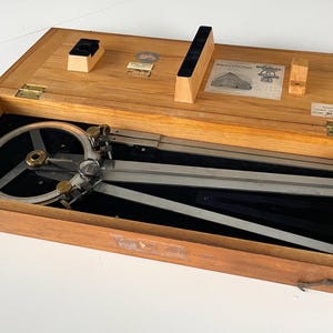 May include: Wooden box containing a drafting instrument with a circular protractor and metal arms. The box is open, revealing the instrument and several wooden and metal components. The box is made of light-colored wood.