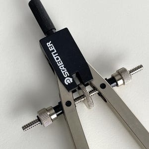 May include: A silver and black Staedtler compass, a drafting tool, is displayed against a white background. The compass features a black adjustment knob and the brand name "Staedtler" printed on the black top. The legs have knurled adjustment screws.