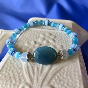 May include: A blue and white beaded bracelet with a large oval blue gemstone in the center. The bracelet has silver accents and is on a white surface.