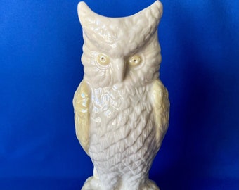 May include: A cream-coloured ceramic owl figurine with yellow highlights. The owl features detailed feather patterns and sits on a base decorated with leaves. The figurine is displayed against a blue backdrop.