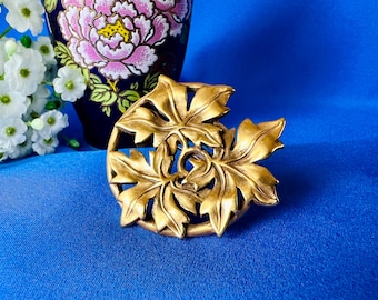 May include: A gold-coloured leaf-shaped brooch with intricate details, set against a blue background. The brooch features a circular design with multiple leaves. A dark blue vase with pink floral patterns is in the background.